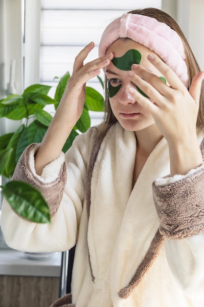 Junges schönes lustiges Mädchen, das Gesichtsmaske Hydrogel grüne Augenklappen anwendet Selbstpflege