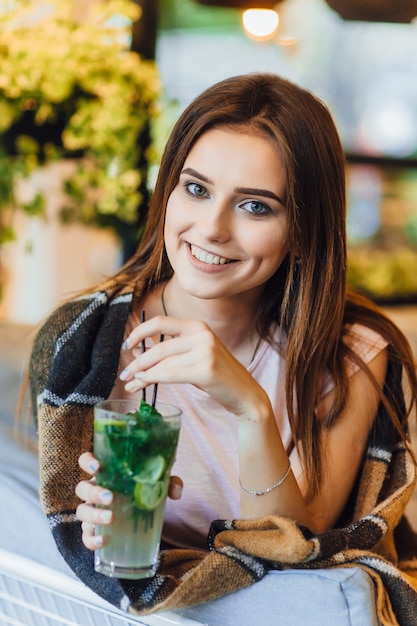 Junges schönes Mädchen auf einer Sommerterrasse in Freizeitkleidung trinkt Cocktail.