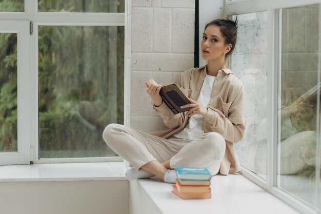 Junges schönes Mädchen, das auf der Fensterbank sitzt und ein Buch liest