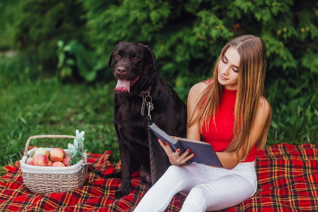 Junges schönes Mädchen, das im Plaid mit ihrem schwarzen Hund labrador im Park sitzt.