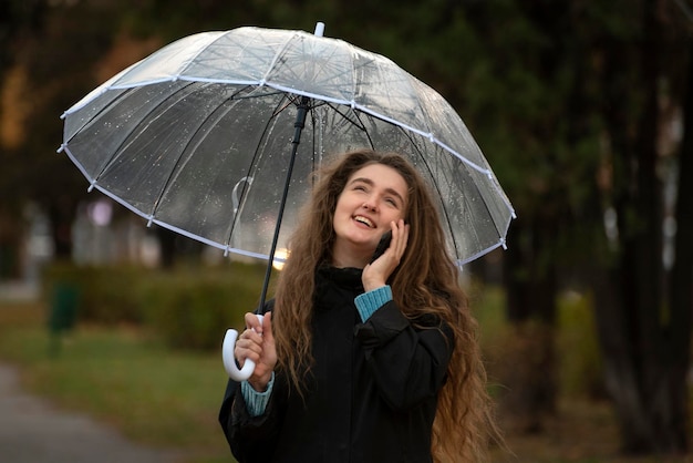 Junges schönes Mädchen im Herbstpark hält transparenten Regenschirm, der am Telefon spricht und lacht