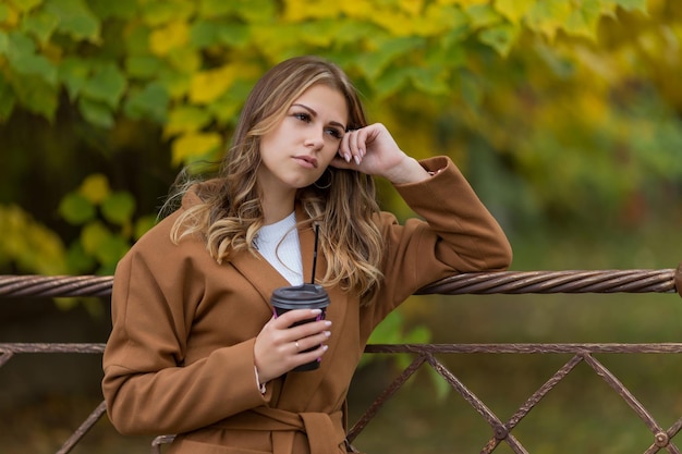 Junges schönes Mädchen in einem braunen Mantel mit einer Tasse Kaffee im Herbstpark Hochwertiges Foto