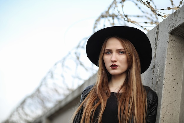 Junges schönes Mädchen in einem Hut und mit einem dunklen Make-up draußen. Mädchen im gotischen Stil auf der Straße. Ein Mädchen geht in einer Lederweste mit Telefon die Straße entlang.