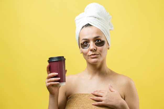 Junges schönes Mädchen in einem weißen Handtuch auf dem Kopf trägt Kollagen-Gel-Patches unter den Augen Maske unter dem Augenbehandlungsgesicht Das Mädchen hält ein Glas Kaffee in den Händen