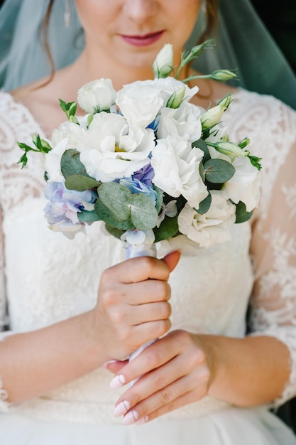 Junges schönes Mädchen in elegantem Kleid steht und hält Handstrauß aus pastellfarbenen Blumen und Grüns mit Band in der Natur Die Braut hält einen Hochzeitsstrauß im Freien