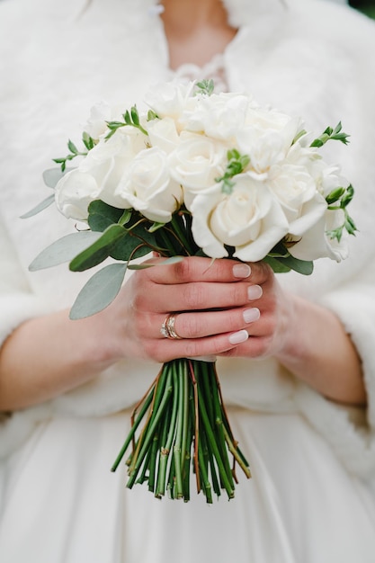 Junges schönes Mädchen in elegantem Kleid steht und hält Handstrauß aus weißen Blumen und Grüns mit Band in der Natur Die Braut hält einen Hochzeitsstrauß im Freien