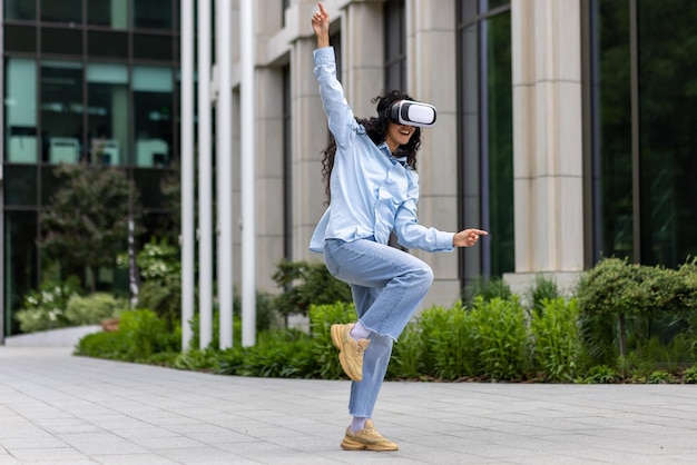 Junges schönes Mädchen in Hemd und Jeans tanzt vor einem Bürogebäude und trägt eine virtuelle Realität
