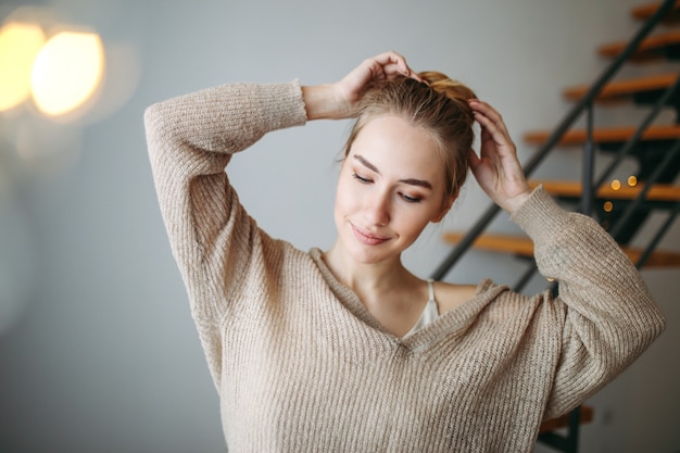 Junges schönes Mädchen mit dem blonden Haar, wenn zu Hause beige Seidenkleid und gemütliche Wolljacke vor Treppe geglättet werden