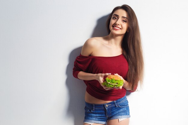 Junges schönes Mädchen mit einem Burger in ihren Händen. Studioaufnahme. Modell. Fast-Food-Mahlzeit. Mädchen in Shorts und einem T-Shirt, Sonnenbrille im Gesicht. Das Konzept des Essens.