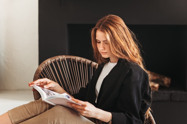 Junges schönes Mädchen mit langen Haaren liest ein Buch, das auf einem Stuhl im Wohnzimmer sitzt