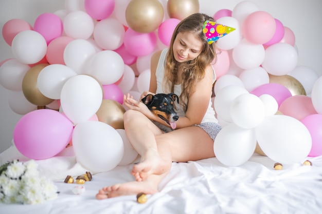 Junges schönes Mädchen wachte umgeben von Luftballons am Geburtstag mit ihrem Haustierhund auf