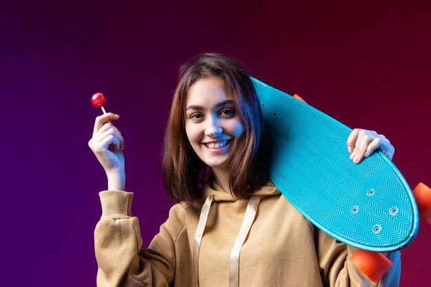 Junges schönes modisches Hipster-Mädchen in einem Hoodie hält ein Skateboard in ihren Händen auf einem violetten Studiohintergrund Isst einen Lollipop