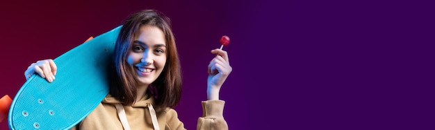 Junges schönes modisches Hipster-Mädchen in einem Hoodie hält ein Skateboard in ihren Händen auf einem violetten Studiohintergrund Isst einen Lollipop