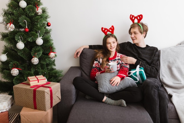 Junges schönes Paar, das zu Hause auf dem Sofa sitzt und Hirschhörner trägt, während es Zeit zusammen mit dem Weihnachtsbaum in der Nähe verbringt