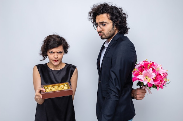 Junges schönes Paar verwirrter Mann, der Blumenstrauß hinter seinem Rücken versteckt und seine verärgerte Freundin mit Schachtel Pralinen betrachtet, die internationalen Frauentag 8. März feiern