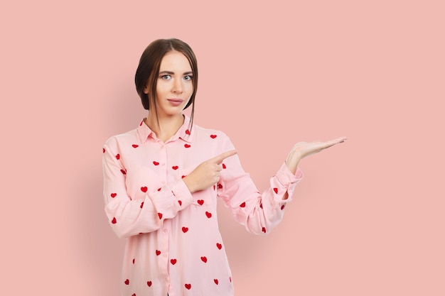 Junges schönes ruhiges Mädchen auf einem rosa Hintergrund in einem rosa Hemd mit roten Herzen zeigt mit den Fingern auf den Ort für Werbeangebote oder Werbung