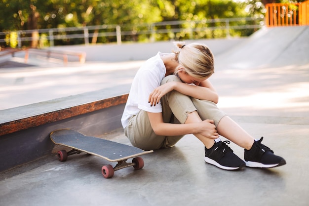 Junges Skatermädchen, das ihr schmerzhaftes Bein hält und mit Skateboard in der Nähe des isolierten Skateparks weint