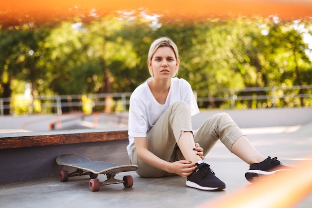 Junges skatermädchen schaut traurig in die kamera und hält ihr schmerzhaftes bein mit skateboard in der nähe, während sie zeit im skatepark verbringt