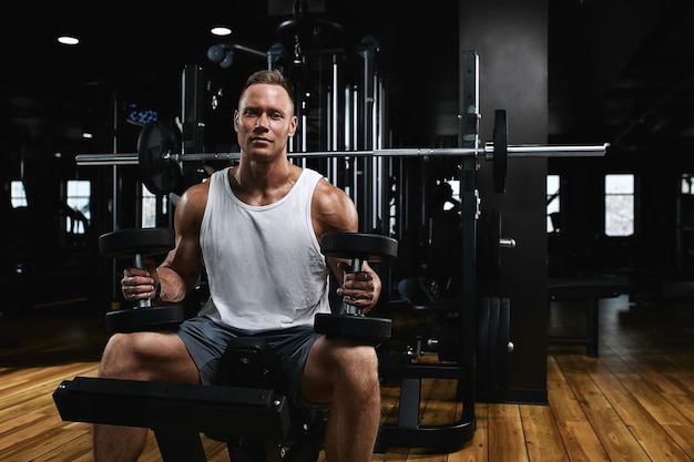 Junges Sportathleten-Fitness-Modell in der Turnhalle schüttelt seinen Bizeps mit Hanteln. Sportliche Motivation, zurückhaltend, hoher Kontrast. Das Konzept eines gesunden Lebensstils, Lebensbewegung, Kopienraum.