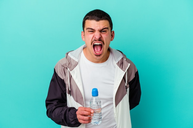 Junges sportliches kaukasisches Manntrinkwasser isoliert auf Blau, das sehr wütend und aggressiv schreit.