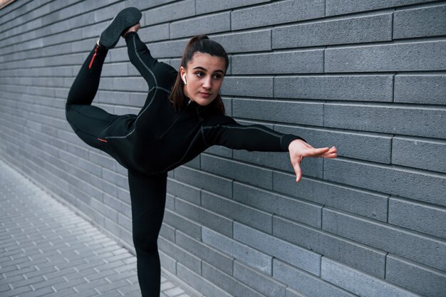 Junges sportliches Mädchen in schwarzer Sportbekleidung im Freien, das Yoga-Übungen in der Nähe einer grauen Wand macht
