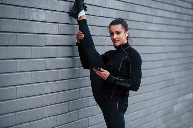 Junges sportliches Mädchen mit Telefon in schwarzer Sportbekleidung, das die Beine mit grauer Wand im Freien streckt