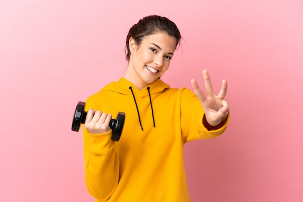 Junges Sportmädchen, das Gewichtheben über lokalisiertem rosa Hintergrund glücklich macht und drei mit den Fingern zählt