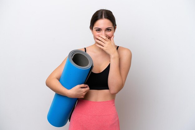 Junges Sportmädchen, das zu Yoga-Kursen geht, während es eine Matte isoliert auf weißem Hintergrund hält, glücklich und lächelnd, den Mund mit der Hand bedeckend