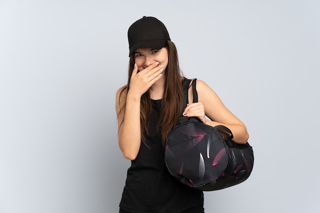 Junges Sportmädchen mit Sporttasche lokalisiert auf grauem glücklichem und lächelndem bedeckendem Mund mit Hand