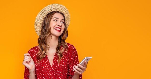 Junges stilvolles Mädchen in einem roten Vintage-Kleid, ein Strohhut mit rotem Lippenstift lächelt, hört Musik auf dem Handy und betrachtet den Kopierbereich, der auf einem gelben und orangefarbenen Hintergrund isoliert ist