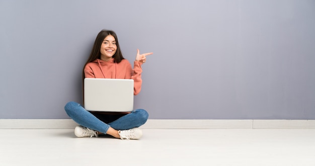 Junges Studentenmädchen mit einem Laptop auf dem Boden Finger auf die Seite zeigend