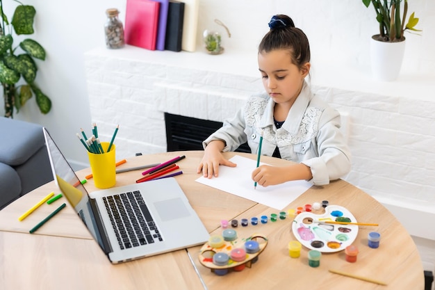 Junges süßes Mädchen, das eine Zeit damit verbringt, mit Online-Lektionen auf dem Laptop zu zeichnen. Online-Bildung im Fernstudium.