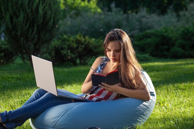 Junges süßes Mädchen mit Laptop, sitzen auf Sitzsack im Garten oder Park, auf grünem Gras. Modernes Lifestyle-Konzept