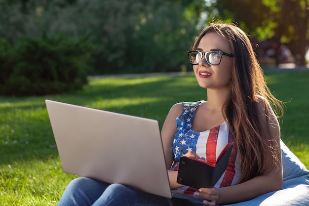 Junges süßes Mädchen mit Laptop, sitzen auf Sitzsack im Garten oder Park, auf grünem Gras. Modernes Lifestyle-Konzept