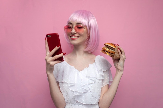 Junges süßes Mädchen mit rosa kurzen Haaren, das Burger hält und Smartphone glückliche Frau mit Telefon verwendet