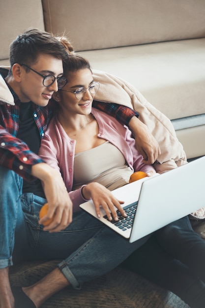 Junges süßes Paar, das auf dem Boden nahe der Couch sitzt, während sie etwas auf einem Laptop beobachtet und Orangen isst, die mit einer Steppdecke bedeckt sind