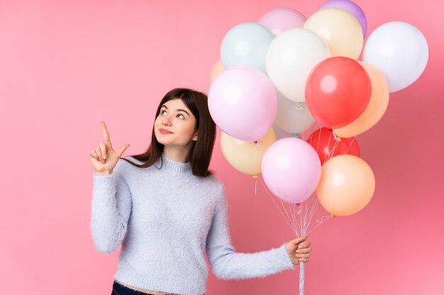 Junges Teenager-Mädchen, das viele Luftballons über rosa Wand hält, die mit dem Zeigefinger eine große Idee zeigt