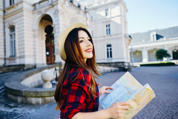 Junges Touristenmädchen mit braunem Haar, das Hut und rotes Hemd trägt, Karte am alten europäischen Stadthintergrund hält und lächelt, reist.