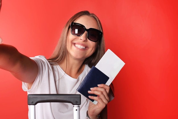 Junges touristisches Mädchen in Freizeitkleidung des Sommers, mit Sonnenbrille, rotem Koffer, Reisepass lokalisiert auf rotem Hintergrund.