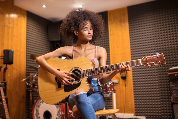 Junges trendiges schwarzes Mädchen mit dem langen lockigen Haar, das Gitarre spielend in einem Aufnahmestudio mit einem Lächeln sitzt