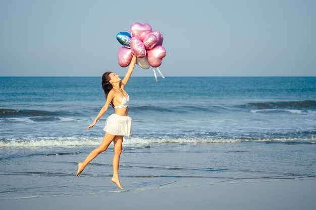 Junges und schönes Mädchen, das Heliumballons hält, vor Freude am Meer läuft und springt. Urlaub und Urlaubskonzept.