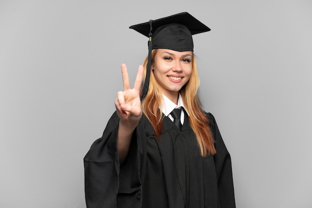 Junges Universitätsabsolventmädchen über isoliertem Hintergrund, der lächelt und Siegeszeichen zeigt