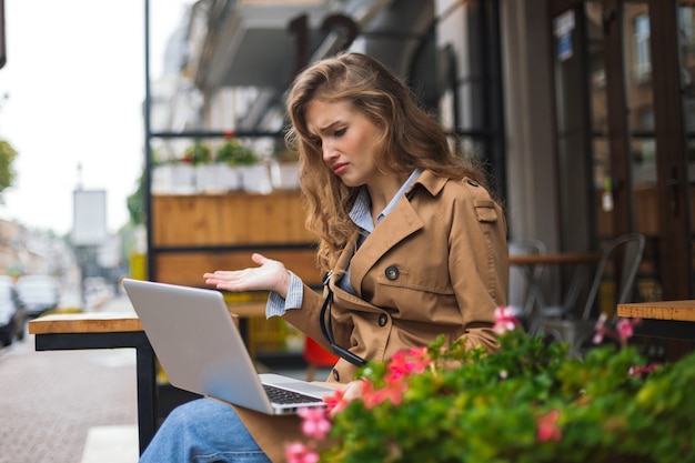 Junges verärgertes Mädchen im Trenchcoat, das traurig am Laptop arbeitet, während Zeit im Freien an der Caféterrasse verbringt