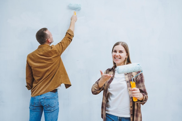 Junges verliebtes Ehepaar in Hemden und Jeans, das Renovierungsarbeiten durchführt, um Wände mit einer Walze zu erneuern und sich auf den Umzug in ein neues Haus mit selektivem Fokus vorbereitet