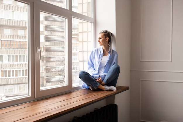 Junges verträumtes Mädchen mit Dreadlocks, die auf einem Fenster sitzen