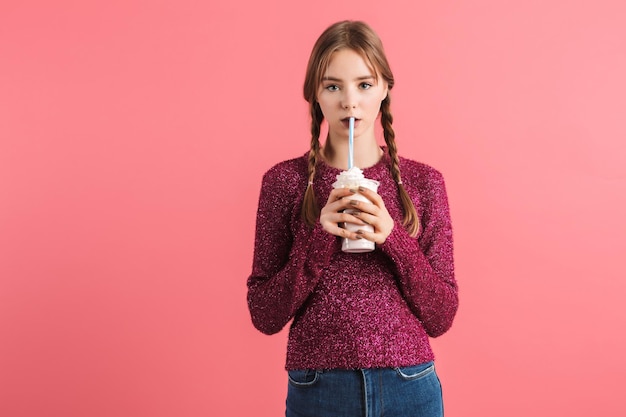 Junges verträumtes Mädchen mit zwei Zöpfen in Pullover und Jeans, das Milchshake trinkt und nachdenklich in die Kamera schaut, isoliert auf rosafarbenem Hintergrund