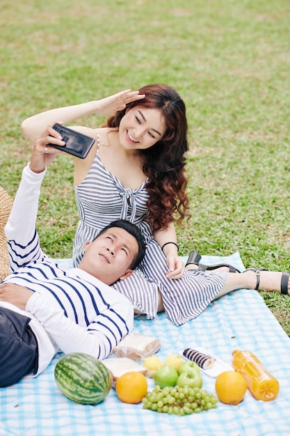 Junges vietnamesisches Paar, das ein romantisches Picknick im Stadtpark genießt und zusammen Selfie macht