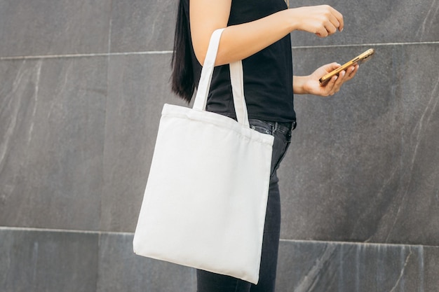 Junges vorbildliches Mädchen auf der Straße mit weißer Öko-Tasche und Handy-Attrappe