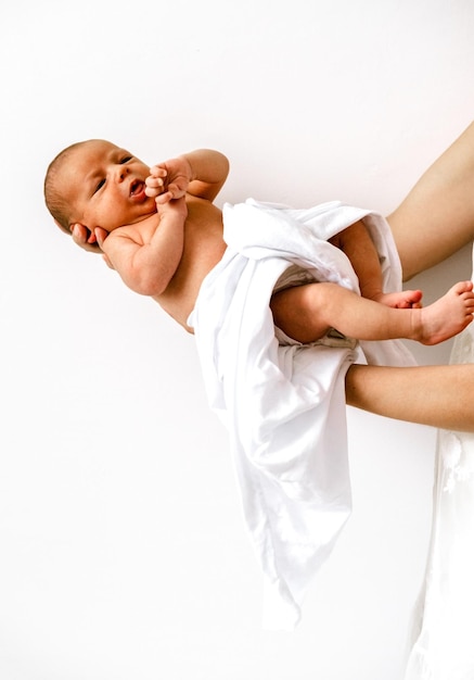 Junges weinendes Baby, bedeckt mit weißer Decke in weiblichen Händen auf weißem Wandhintergrund
