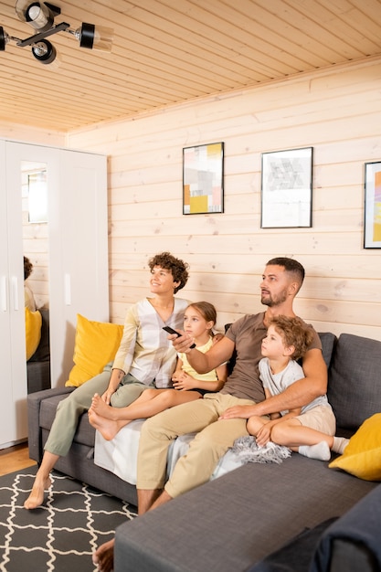Junges zeitgenössisches Paar und ihre zwei süßen kleinen Kinder in Freizeitkleidung sitzen auf der Couch im Wohnzimmer und sehen am Wochenende Fernsehsendungen?